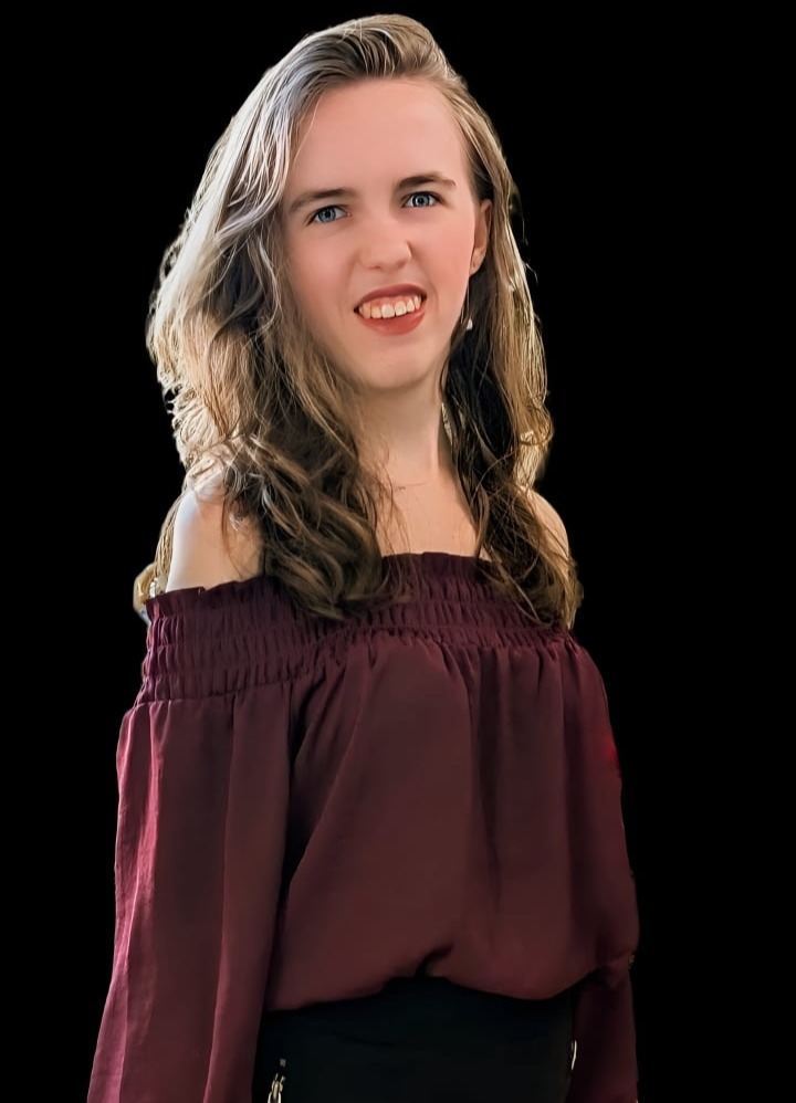 Stefanie, a blonde-haired, blue-eyed woman wearing a burgundy blouse, standing against a black background.